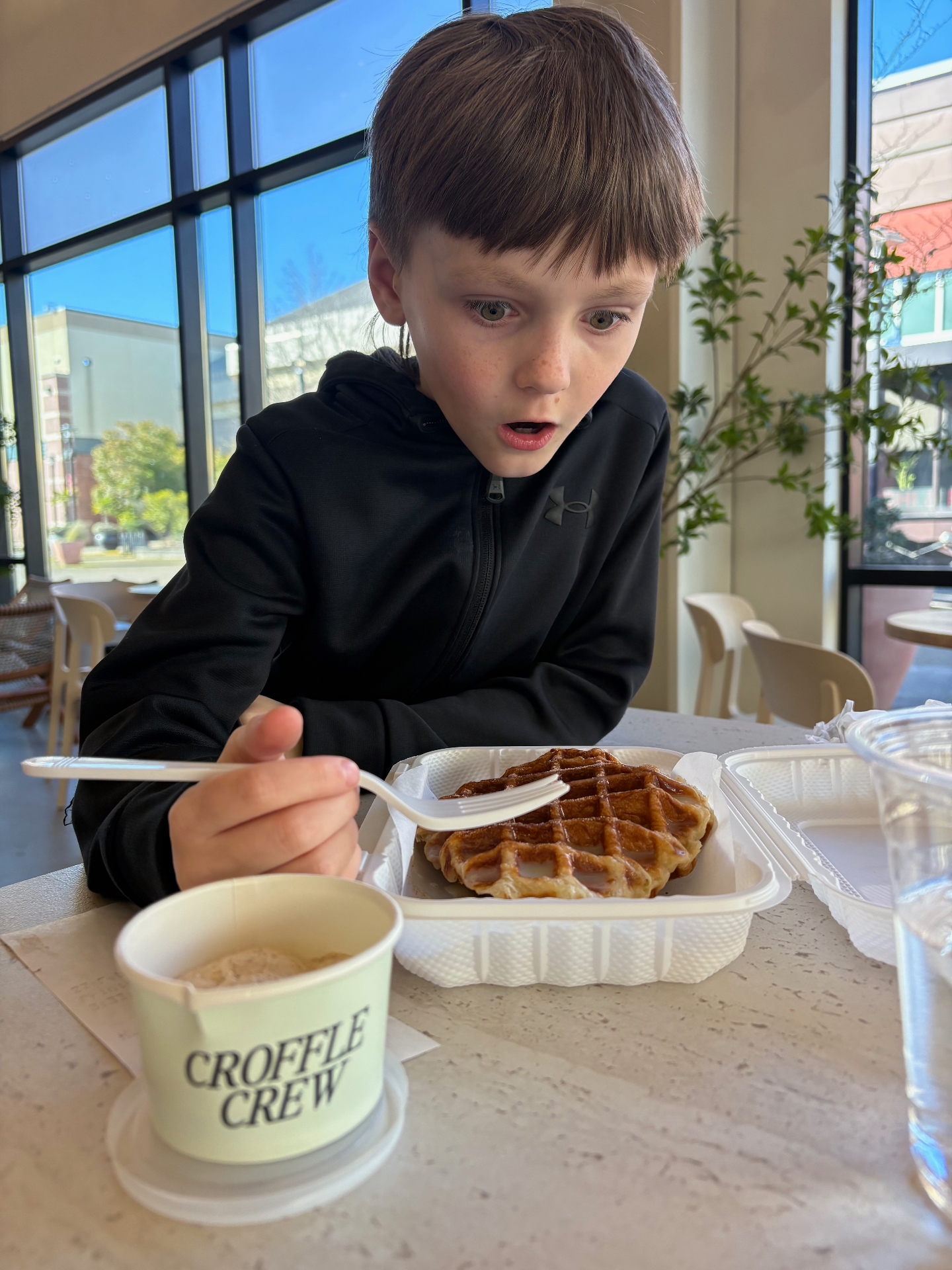 Younger son sitting at a sunlit cafe counter, mid-bite of a fresh croffle.
